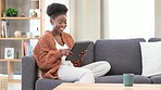 Woman laughing and browsing social media online on a digital tablet while relaxing on the couch at home. One young black female smiling while searching the internet and texting friends on the web