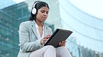 Serious business woman typing on a tablet and listening to music using headphones while sitting outside a building. Female corporate professional browsing the internet or writing an email outdoors