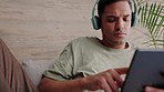 Headphones music, tablet and relax man on sofa with home internet or wifi browsing social media app, website or searching online content. Young guy with wireless technology listening to podcast audio