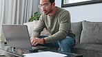 Man, glasses and laptop working on sofa in the living room typing email for business idea at home. Smart male freelancer or typist focused at work on computer sitting on couch planning design ideas