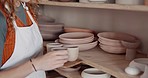 Pottery, creative woman and shelf with clay art in store, workshop and workplace for customers. Female entrepreneur, prepare artistic mold or enjoy sculpture ceramic cups for craft class in studio.