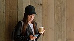 A young woman drinking coffee and reading text messages on her cellphone in front of a wood wall before going to university. A young student reading news on her cellphone and drinking coffee