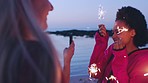 Woman, friends and beach with sparks at night, celebration and happiness outdoor. Young people, phone and New Years on vacation for summer break, travelling for adventure and fireworks on holiday.