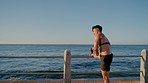 Fitness, exercise and man stretching outdoor at sea promenade while listening to music for workout motivation and energy. Asian athlete runner with ocean and sky horizon for cardio health training