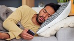 Sick guy using a phone wrapped in a blanket lying on the sofa suffering from fever and heavy coughing. Young male with flu feeling unwell while texting on an online app and browsing social media
