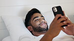 Man, phone and smile lying on bed for social media, chatting or texting while relaxing at home. Happy relaxed male smiling on smartphone in bedroom browsing, streaming or enjoying entertainment