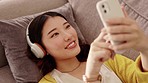 Happy woman, headphones and phone in a living room with social media, music and texting. Home, sofa and smile of a Asian female and gen z student feeling relax from audio and web podcast on couch