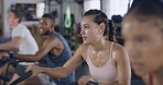Focused and sporty woman during a cycling class at the gym. Fit people using air bikes for cardio exercise in a fitness facility. Group of sweaty men and women listening to music while exercising