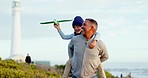 Senior man, child and beach with plane, game or outdoor for love, care or bonding by lighthouse. Grandfather, girl kid and airplane toys on vacation, holiday and play together with piggy back by sea