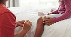 First aid, injury and mom with a bandage for girl in bedroom with care for wound from accident or hurt knee. Mother, nursing or helping child on bed with pain, bruise or support in house or home