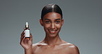 Beauty, face serum and portrait of a woman with bottle cosmetics, smile and dermatology. Natural Indian model person with facial care, skin glow and oil product in hand on grey background in studio