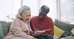 Assisted living, photo album or memory with a senior and black woman caregiver on a living room sofa. Smile, funny and elderly resident looking at a picture book with a nurse in a retirement home