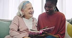 Photo album, assisted living and an old woman laughing with her caregiver on a sofa in the living room. Smile, funny and senior resident looking at a memory with an african nurse in a retirement home