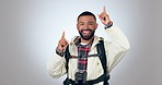 Hiking, travel and face of man in studio pointing up, dance and freedom celebration on grey background. Adventure, portrait and male backpacker dancing for news, announcement or explore opportunity