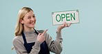 Open sign, woman pointing and face with shop, entrepreneur and startup poster in a studio. Blue background, female person and job portrait with a smile and notice of a retail store or restaurant