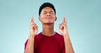 Praying, face and man with fingers crossed in studio for hope, good luck or feedback results on blue background. Help, prayer and male model show hand emoji for waiting, review or lotto prize outcome