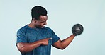 Fitness, man and pointing to bicep with dumbbell for training, exercise or workout in studio on blue background. Weightlifting, person and personal trainer for wellness and healthy body with smile