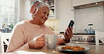 Senior woman, home and headphones with phone, coffee and funny video call, social media or streaming comedy. Elderly person laughing, happy and listening to mobile podcast or meme with tea in kitchen