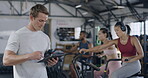 Gym trainer using digital tablet to monitor his HIIT group fitness class. Fit, athletic and active coach browsing health data while diverse people exercise and workout on air bikes in the background