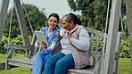 Happy woman, nurse and patient in relax on tablet, bench or swing for entertainment in nature or park. Female person or medical caregiver sitting outdoor with senior, technology or coffee for support