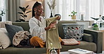 Laundry, housework and a black woman folding washing on a sofa in the living room of her home to tidy. Smile, relax and a happy young housewife cleaning her apartment for housekeeping or hygiene