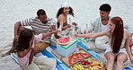 Friends, beach picnic and toast drink for celebration, birthday or new years with food, relax or vacation. Men, women and happy with cheers, bottle or alcohol by ocean, holiday or summer in Cape Town