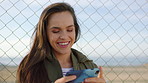 A smiling young woman on holiday in front of a fence watching videos on her cellphone. A young woman laughing while watching videos on her smartphone and standing in front of a fence.