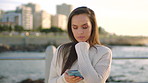 A young woman reading text messages on her smartphone standing on the promenade. A young woman on holiday at the beach reading texts on her cellphone. A woman using her smartphone at the beach