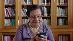 Elderly woman on her phone in a library on the internet doing research or typing a message. Senior librarian networking on social media or checking her emails at work while looking outside.