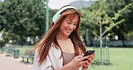 Woman, cellphone and headphones or walking in park or smile or online communication, streaming or playlist. Female person, smartphone and texting in nature or relax weekend or podcast, track or song