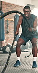 Black man, fitness and battle rope at gym for workout, strength or muscle endurance on floor. Serious African male person, bodybuilder or athlete in sweat, exercise or intense power at health club