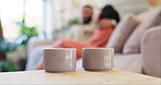 Coffee, steam and cups on table in home for breakfast, latte or cappuccino in the morning closeup. Tea, mug and vapor of drink, beverage or hot espresso on counter in living room with couple on sofa