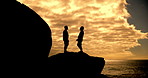 Happy, people and silhouette with sunset, sky and orange clouds for relax, peace and freedom. Friends, shadow and ocean wave for hike adventure together with support and bonding on rocks for wellness