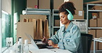 Woman, laptop and headphones at shipping startup, typing or reading by boxes on shelf for supply chain. African person, employee and pc for music, dance or storage with invoice admin for distribution