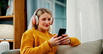 Happy woman, typing and phone with headphones on sofa for online streaming, browsing or connection at home. Female person with headset, smile or scrolling on mobile smartphone for music list or app