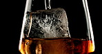 Glass, ice and pour brandy in studio for enjoyment, refreshing and cold beverage. Frozen cube, tumbler and whiskey flow on black background with liquor, drink and aesthetic for testing in distillery