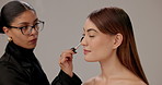 Makeup artist, work and women with brush in studio for cosmetic, beauty and preparing for photoshoot. Beautician, application and people with service for shine, glow and glamour on gray background