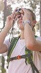 Woman, hiker and sightseeing with binoculars in woods for birdwatching, tourism or nature hobby. Ornithologist, person or traveler with lens for zoom, ecology research or scenery with forest holiday