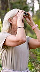 Mature woman, hiker and sightseeing with binoculars in forest for birdwatching, tourism or retirement hobby. Ornithologist, person or traveler with woods for holiday, ecology research or scenery