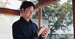 Window, phone and Asian man in home online for internet, connection and networking. Living room, house and Japanese  person on smartphone for website, social media and reading news blog in morning