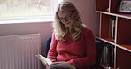 Woman, book and reading to relax, study or learning in home for knowledge, story or information. Senior, happy and studying in house, room or home office for by shelf, books and sitting for education