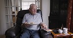 Senior man, eating bread and read magazine, relax and sofa in living room, retirement life and snack in home. Elderly male reading a book, eating food and relaxing on the couch in the lounge in home