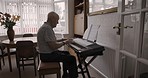 Senior man playing music on a keyboard with sheets in living room for entertainment in his house. Music, hobby and elderly guy in retirement with a song book to play on the piano in lounge at home.