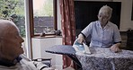 Ironing, laundry and senior couple talking during housework, chores and home cleaning. Communication, speaking and elderly woman with company from a man during housewife work in the living room