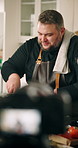 Cooking, live streaming and man in kitchen with food for culinary lesson, meal tutorial and dinner in home. Camera, influencer and person with ingredients for nutrition, gourmet recipe and cuisine