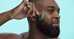 Happy, black man and skincare with jade roller for anti aging or facial treatment on a blue studio background. Male person, model or smile with beauty tool for spa, face massage or lymphatic drainage