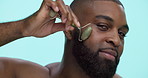 Happy, black man and face with jade roller for anti aging treatment on a blue studio background. Portrait, male person or model with beauty tool or stone for spa, facial massage or lymphatic drainage