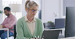A mature business woman wearing spectacles looking annoyed while working in a office on a laptop and sitting at a desk 