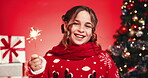 Christmas tree, face and sparkler with girl on red background for celebration of holiday season. December, fireworks and smile of happy child in studio for excitement of fitness gifts or presents