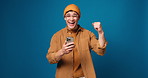 Happy man, face and winning with phone in studio for good news or bonus on a blue background. Portrait, male person or model with smile, mobile smartphone or fist pump for success on mockup space